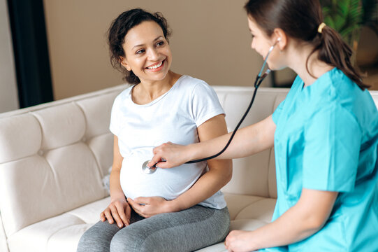 Happy pregnant woman and female doctor. Friendly doctor examines pregnant mixed race smiling woman, listening to baby's heartbeat, sitting on a sofa at home or hospital,healthy pregnancy concept