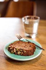 A slice of banana bread with pumpkin seeds on a blue plate, a dessert fork, a glass of water. Wooden table, cafe.