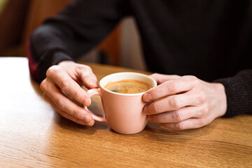 Man holding a pink ceramic cup of hot black coffee in his hands, sitting at the wooden table in a cafe.