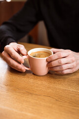 Man holding a pink ceramic cup of hot black coffee in his hands, sitting at the wooden table in a cafe. Copy space.