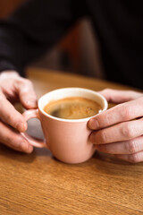 Man holding a pink ceramic cup of hot black coffee in his hands, sitting at the wooden table in a cafe. Close-up photo