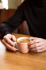 Man holding a pink ceramic cup of hot black coffee in his hands, sitting at the wooden table in a cafe.