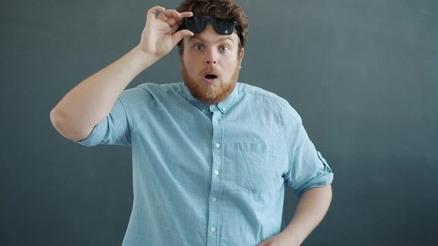 Portrait of shocked guy looking at camera then taking off sunglasses standing on gray background alone with astonished face. People and emotions concept.