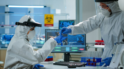 Biologist doctor in ppe suit checking liquid sample typing on pc in equipped laboratory. Team examining vaccine evolution in medical lab using high tech and chemistry tools for scientific research