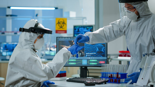 Biologist doctor in ppe suit checking liquid sample typing on pc in equipped laboratory. Team examining vaccine evolution in medical lab using high tech and chemistry tools for scientific research