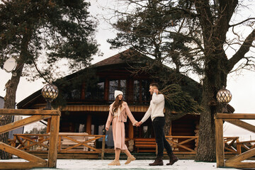 Young beautiful happy lovers man and woman, love story in winter with a live Christmas tree in their hands on the background of a wooden house