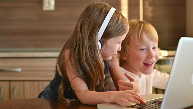 Excited ittle girl and boy using a laptop with wireless headphones and having fun at home. Kids and technology.