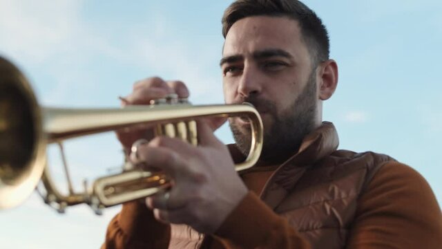 Low angle chest-up POV of young Caucasian man playing trumpet in foreground of blue sky outdoors, looking on camera