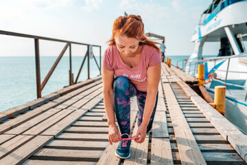 An adult woman in sportswear, sitting down tying her shoelaces. In the background, the pier with the ship and the sea. The concept of sport and a healthy lifestyle