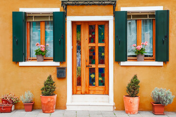 Door and windows with flowers on the yellow facade of the house. Colorful architecture in Burano island, Venice, Italy.