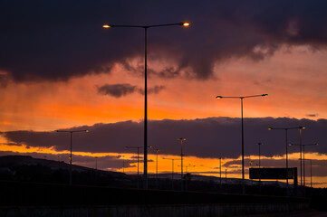 Fototapeta na wymiar Spectacular red sunset with dark rainy clouds over the bridge with silhouettes of people and vehicles and tall street lanterns along highway M50 in Dublin, Ireland. Beautiful cold sunset clouds