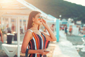 Portrait of a beautiful woman in sunglasses posing on the beach. Sun beds and umbrellas are in the background. Sunlight. The concept of summer luxury holidays