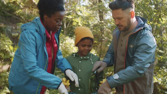 Little mixed race boy holding trash bag while Afro-American mother and Caucasian father collecting garbage into it after staying at campsite in forest