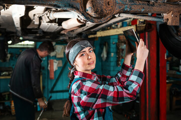 Naklejka na ściany i meble Portrait of a young beautiful female mechanic in uniform who conducts a car inspection using a diagnostic gadget. The car is on the lift
