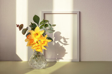 Daffodil bouquet in glass vase and picture frame against white wall in sunlight. Spring concept