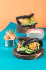Two asian street fast food-Bao Burger with breaded shrimp, fresh cucumbers and seaweed in a black cuttlefish ink bun on a bright blue-orange background.
