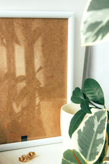 Still life. Cup of coffee, blank beige wooden frame, beautiful gold earrings and indoor evergreen potted plant on the white wall. Scandinavian working space, home office