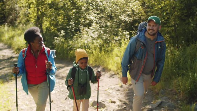 Happy multiethnic parents and little mixed race son smiling and talking while walking in woods with trekking poles and backpacks on summer day