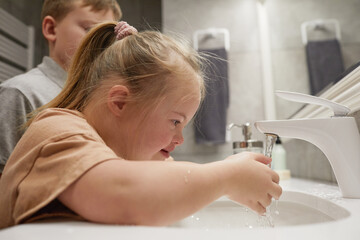 Side view portrait of cute girl with down syndrome washing hands while standing by sink at home,...