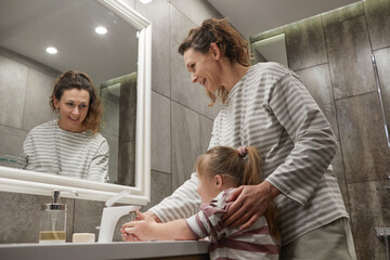 Side view portrait of smiling mother helping little girl with down syndrome wash hands at sink in...