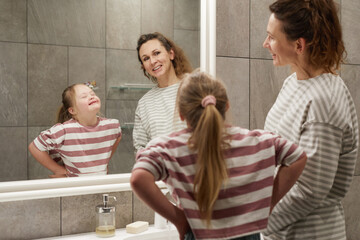 Portrait of cute little girl with down syndrome making faces at mirror while having fun with mother in bathroom, copy space © Seventyfour