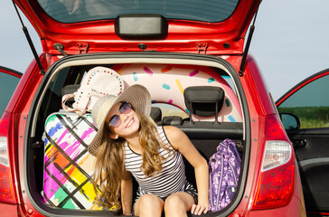 Family ready for the travel on summer vacation. Cute happy smiling little girl child is sitting in the red car with luggage and bags outdoors.