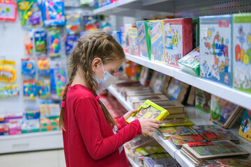  A child in a medical mask is choosing a children's toy in the store. A girl is shopping amid the...
