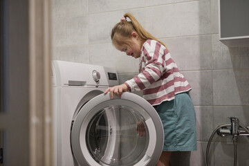 Side view portrait of cute girl with down syndrome using washing machine at home, copy space
