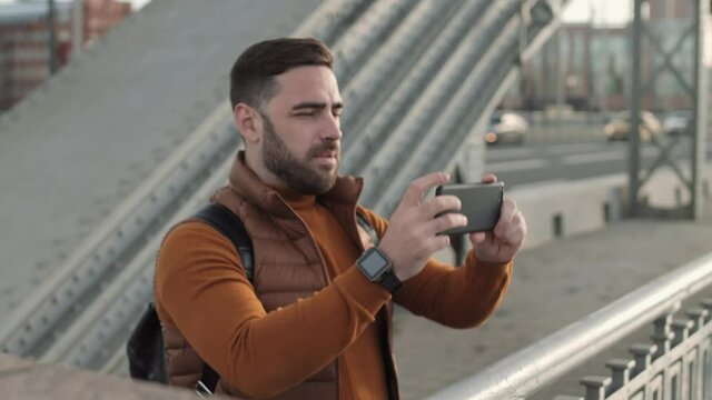 Waist-up shot of young male Caucasian tourist with backpack stopping on bridge, taking pictures with smartphone, then walking away, cars going on background