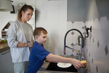 Side view portrait of little boy washing dishes at home while standing by sink in kitchen with happy mother, copy space