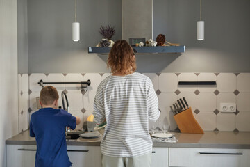 Back view portrait of little boy washing dishes in kitchen while helping mother, copy space