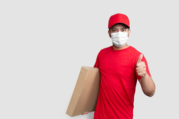 Delivery man employee in red cap blank t-shirt uniform face mask hold empty cardboard box showing thumbs up isolated on white background