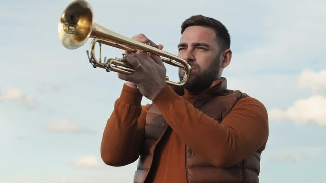 Low angle waist-up of young bearded Caucasian man wearing brown sweater and vest, playing trumpet in foreground on blue sky outdoors