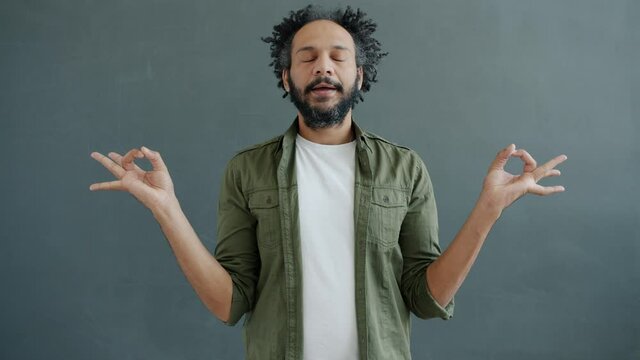 Slow motion portrait of enlightened mixed race man meditating holding hands in namaste gesture standing on gray bacckground with closed eyes relaxing