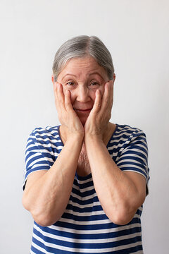 Elderly woman, gray-haired lady of the 60s in a striped white and blue dress shocked, surprised and amazed expression with hands on her face on a white background