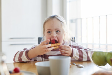 Front view portrait of cute girl with down syndrome eating sweet waffles while enjoying breakfast with family at home, copy space