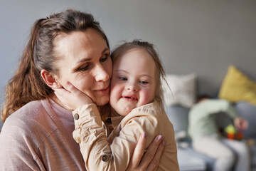 Candid portrait of cute girl with down syndrome hugging mother lovingly in simple home interior,...