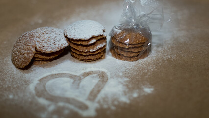 Several cookies, lying on top of each other, are stacked, one stack is wrapped in cellophane wrap. In front, a heart is drawn on white powdered sugar with a finger.