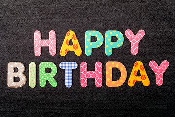 Card with Happy Birthday words made from mixed vivid colored wooden letters on a textured dark black textile material that can be used as a message.