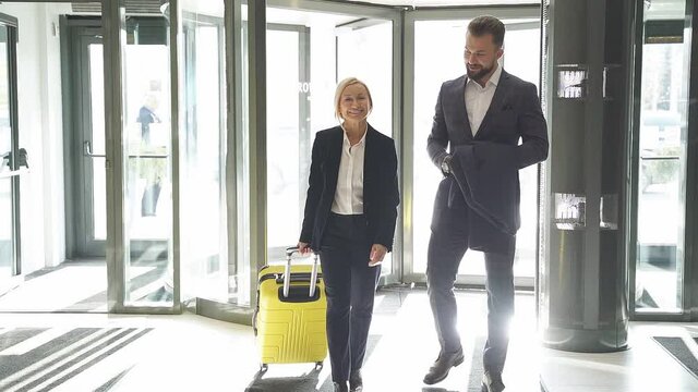 Business woman, accompanied by a manager, enters the hotel with suitcase, in formal wear.