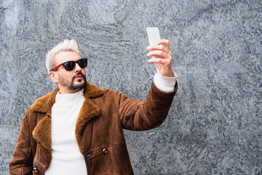 Hipster bearded influencer with platinum blonde hair and sunglasses making selfie with brand new smartphone, fashionable vlogger creating content in outdoor, copy space