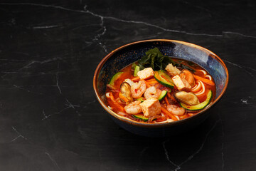 Asian ramen soup with shrimps, mussels, tofu, carrot, zucchini and seaweed. Dish isolated in a dark blue bowl, close-up on a black marble background. Asian cuisine. Copy space.