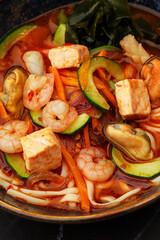 Asian ramen soup with shrimps, mussels, tofu, carrot, zucchini and seaweed. Dish isolated in a dark blue bowl, close-up on a black marble background. Asian cuisine.
