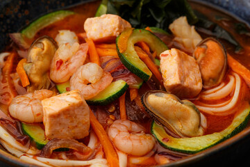 Asian ramen soup with shrimps, mussels, tofu, carrot, zucchini and seaweed. Dish isolated in a dark blue bowl, close-up on a black marble background. Asian cuisine.