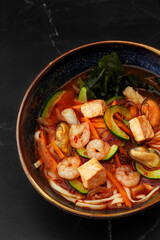 Asian ramen soup with shrimps, mussels, tofu, carrot, zucchini and seaweed. Dish isolated in a dark blue bowl, close-up on a black marble background. Asian cuisine.