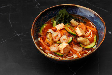Asian ramen soup with shrimps, mussels, tofu, carrot, zucchini and seaweed. Dish isolated in a dark...