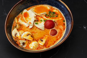 Asian chicken curry soup with chicken breast, tomatoes, mushrooms and spring onion. Close-up photo of the dish served in a dark blue bowl, isolated on a black marble background. Asian cuisine.