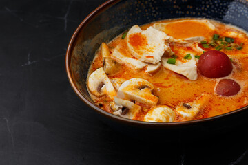 Asian chicken curry soup with chicken breast, tomatoes, mushrooms and spring onion. Close-up photo of the dish served in a dark blue bowl, isolated on a black marble background. Asian cuisine.