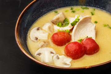 Asian coconut chicken soup with chicken breast, tomatoes, mushrooms and spring onion. Close-up photo of the dish served in a dark blue bowl, isolated on a black marble background. Asian cuisine.