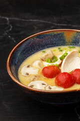 Asian coconut chicken soup with chicken breast, tomatoes, mushrooms and spring onion. Close-up photo of the dish served in a dark blue bowl, isolated on a black marble background. Asian cuisine.